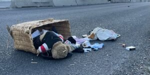 Cesta grande volcada en una carretera vacía, con un oso de peluche y objetos personales regados sobre el asfalto.