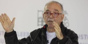 Retrato del periodista José María "Chema" Salcedo hablando frente a un micrófono en una conferencia. Viste una casaca de cuero negra, lentes de marco rojo y gesticula con las manos.